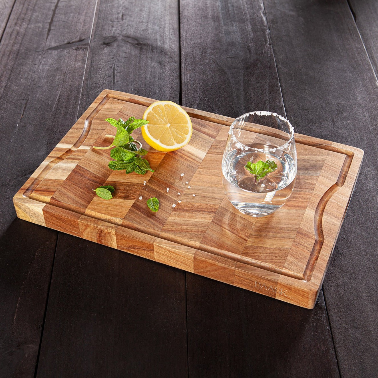 Acacia wood chopping board. Detailed chevron pattern. Built in grove handles with juice groove waith glass of water cut lemon amd mint
