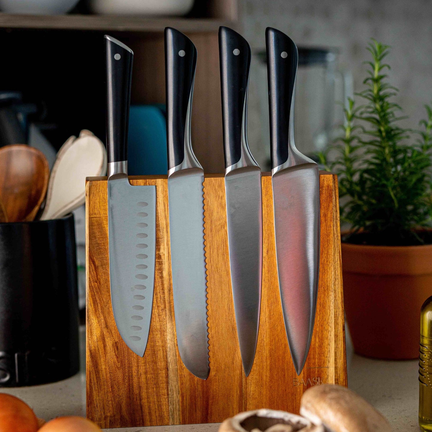 Acacia wood magnetic knife block holding chef knives. On kitchen bench with utensils and potted herbs in background